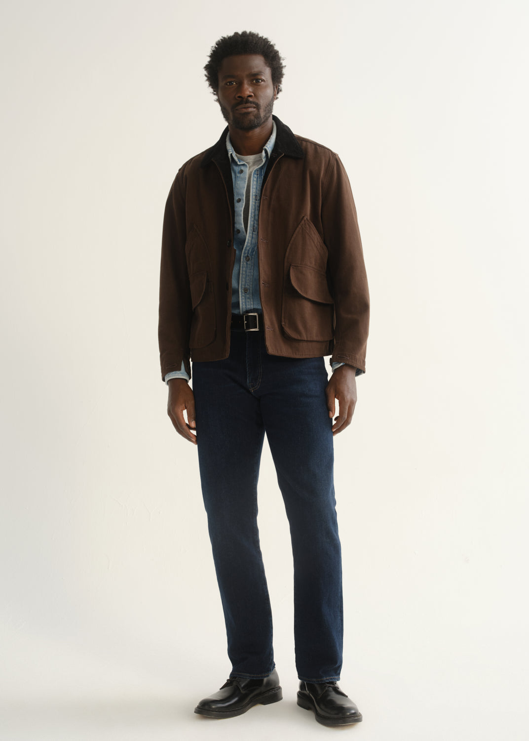 Person wearing dark indigo straight leg jeans with a brown jacket, blue button-up shirt, and black shoes, front full body view on a light grey background.