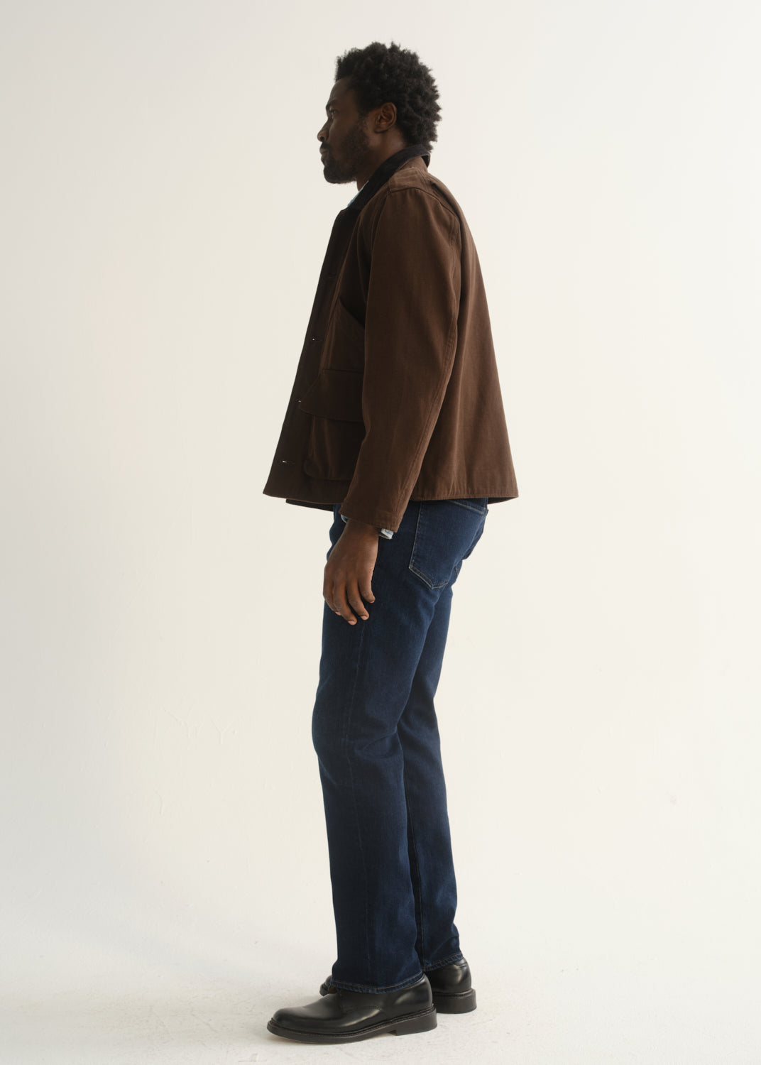 Person wearing dark indigo straight leg jeans with a brown jacket and black shoes, side full body view on a light grey background.