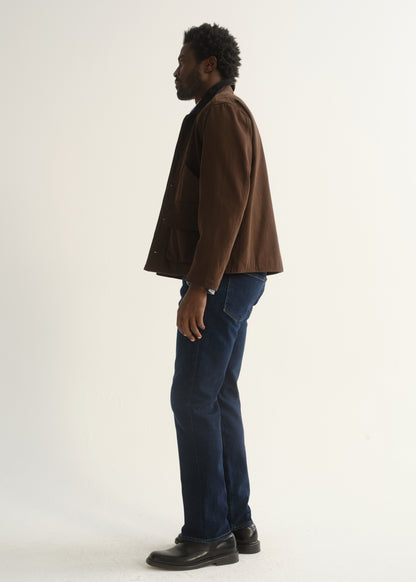 Person wearing dark indigo straight leg jeans with a brown jacket and black shoes, side full body view on a light grey background.