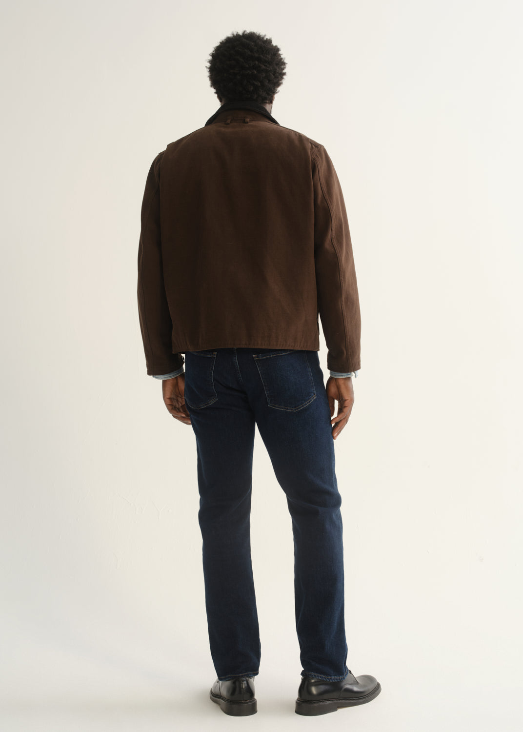 Person wearing dark indigo straight leg jeans with a brown jacket and black shoes, back full body view on a light grey background.