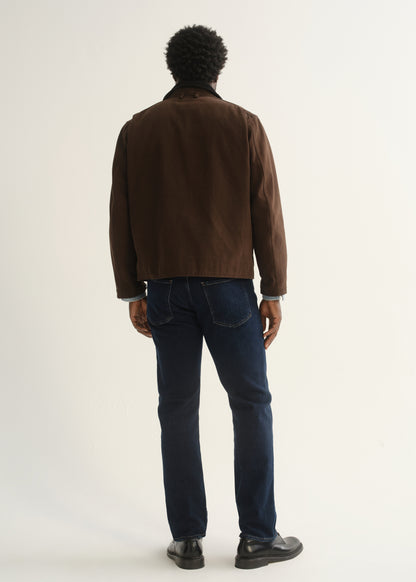 Person wearing dark indigo straight leg jeans with a brown jacket and black shoes, back full body view on a light grey background.