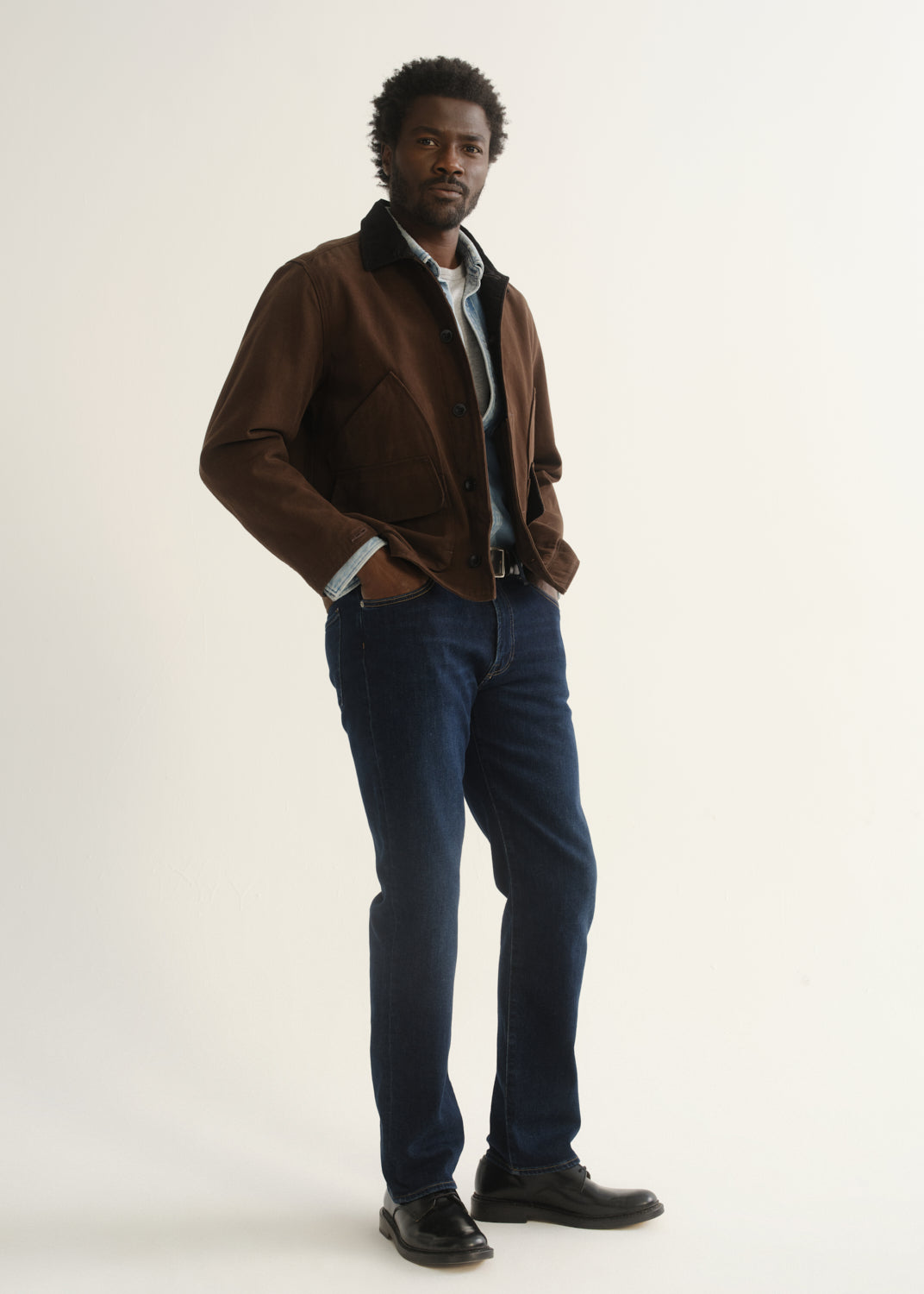 Person wearing dark indigo straight leg jeans with a brown jacket, blue button-up shirt, and black shoes, angled front full body view on a light grey background.
