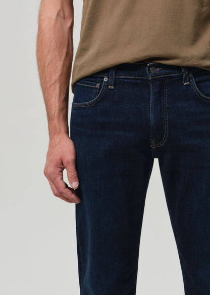 Person wearing dark blue jeans and a brown shirt on a light gray background