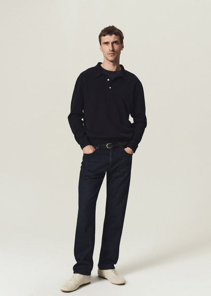 A man modeling a dark navy long-sleeve polo shirt, dark navy trousers, a black belt, and off-white sneakers against a plain light beige studio background.