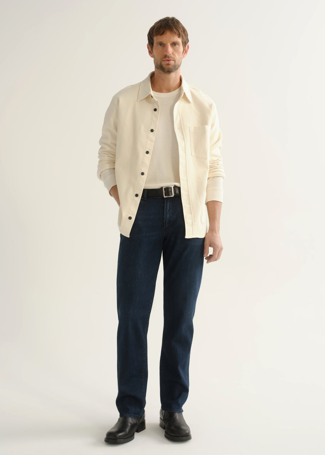 Person wearing dark indigo straight leg jeans with a cream button-up jacket over a grey shirt and black boots, front full body view on a light grey background.
