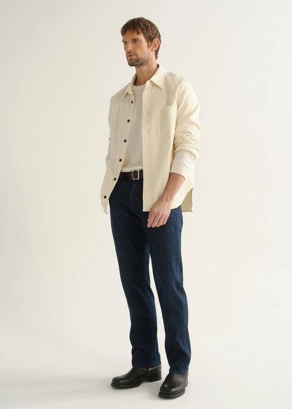 Person wearing dark indigo straight leg jeans with a cream button-up jacket and black boots, angled front full body view on a light grey background.