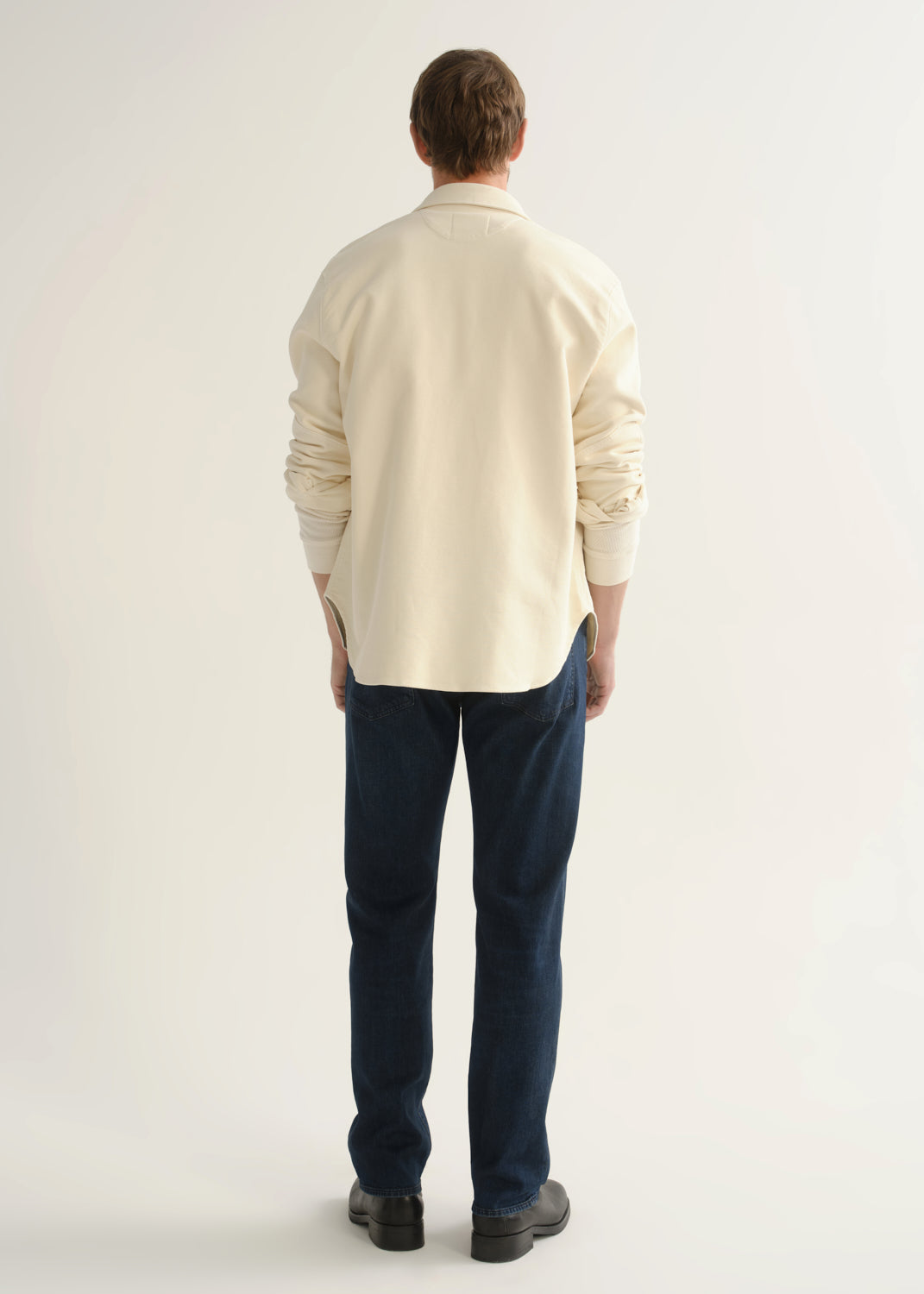 Person wearing dark indigo straight leg jeans with a cream button-up jacket and black boots, back full body view on a light grey background.