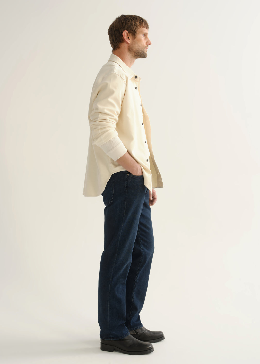 Person wearing dark indigo straight leg jeans with a cream button-up jacket and black boots, side full body view on a light grey background.
