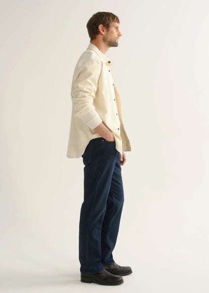 Person wearing dark indigo straight leg jeans with a cream button-up jacket and black boots, side full body view on a light grey background.