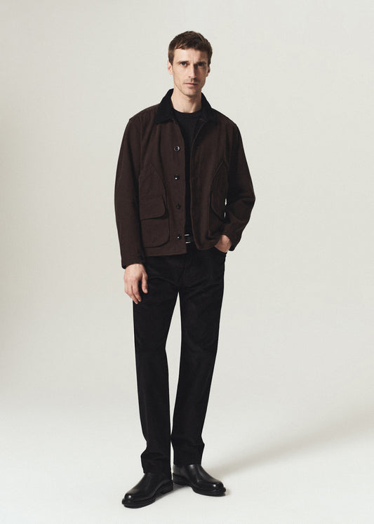 A man modeling a dark brown corduroy jacket with a black collar, worn over a black shirt and black trousers, standing against a light gray studio background.