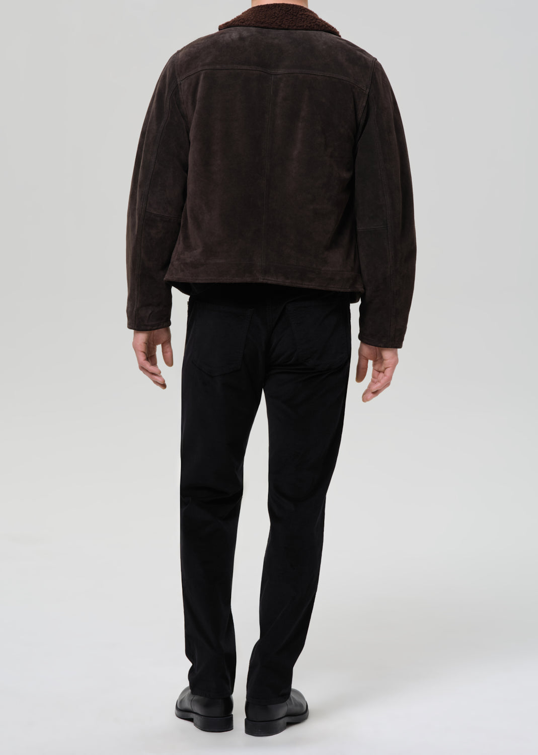 Person wearing a brown jacket and black pants on a grey background