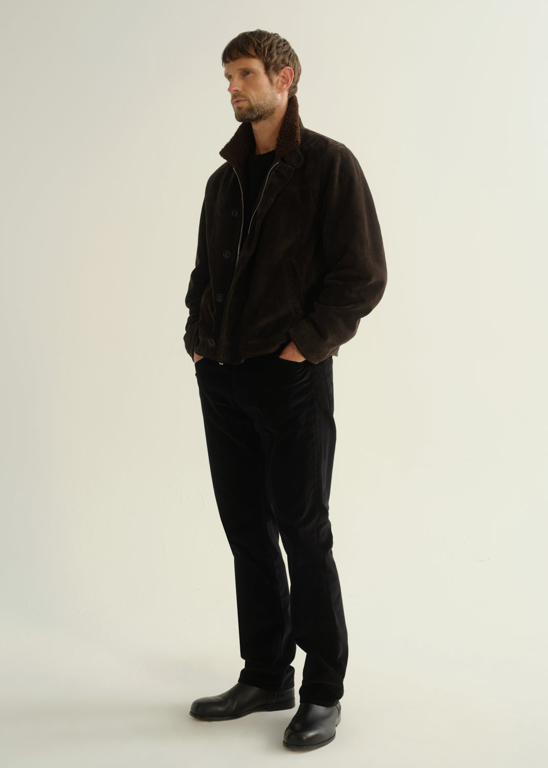 Person wearing black straight leg jeans with a dark brown jacket and black boots, angled front full body view on a light grey background.