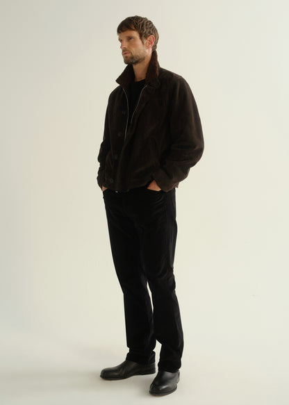 Person wearing black straight leg jeans with a dark brown jacket and black boots, angled front full body view on a light grey background.