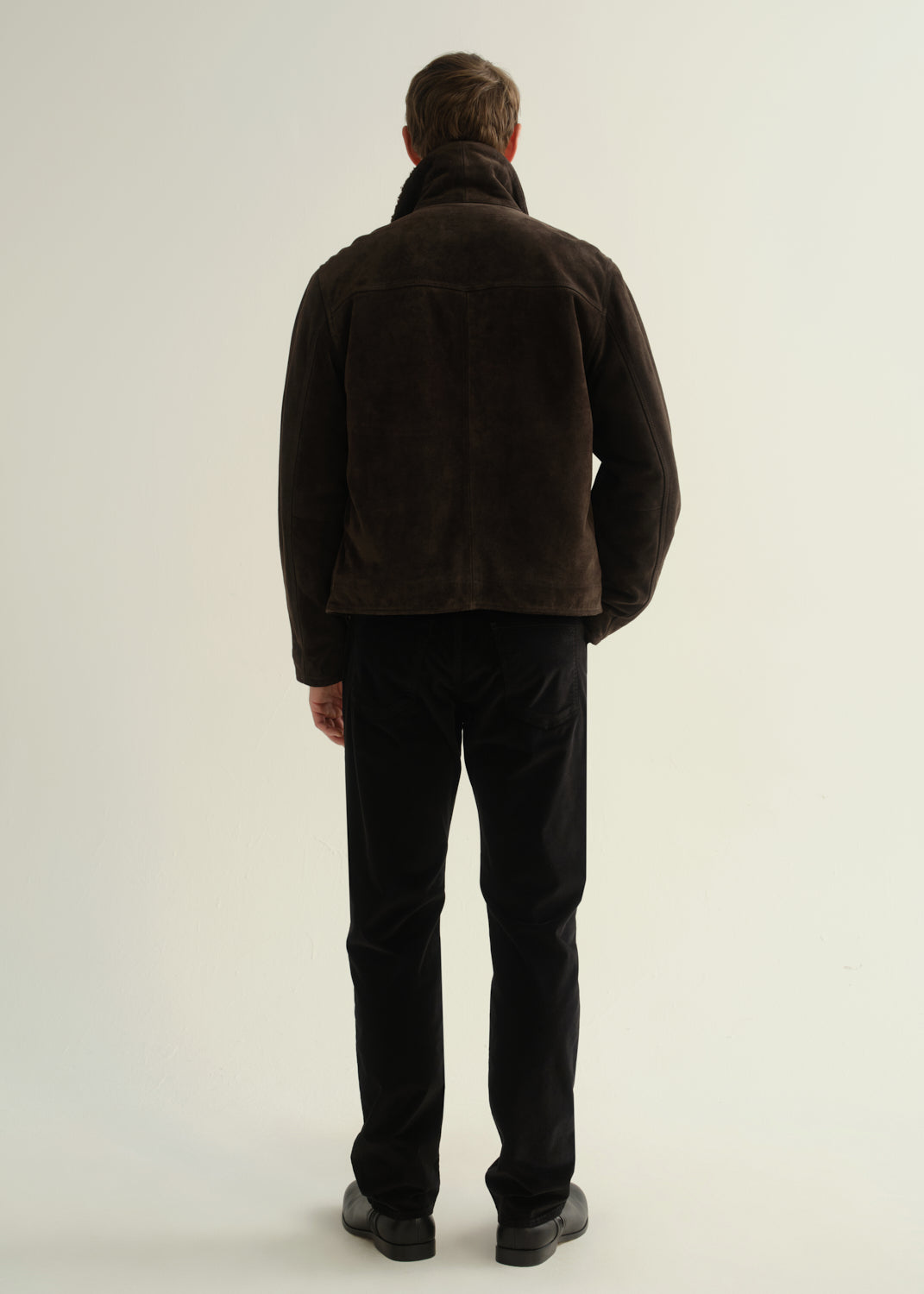 Person wearing black straight leg jeans with a dark brown jacket and black boots, back full body view on a light grey background.
