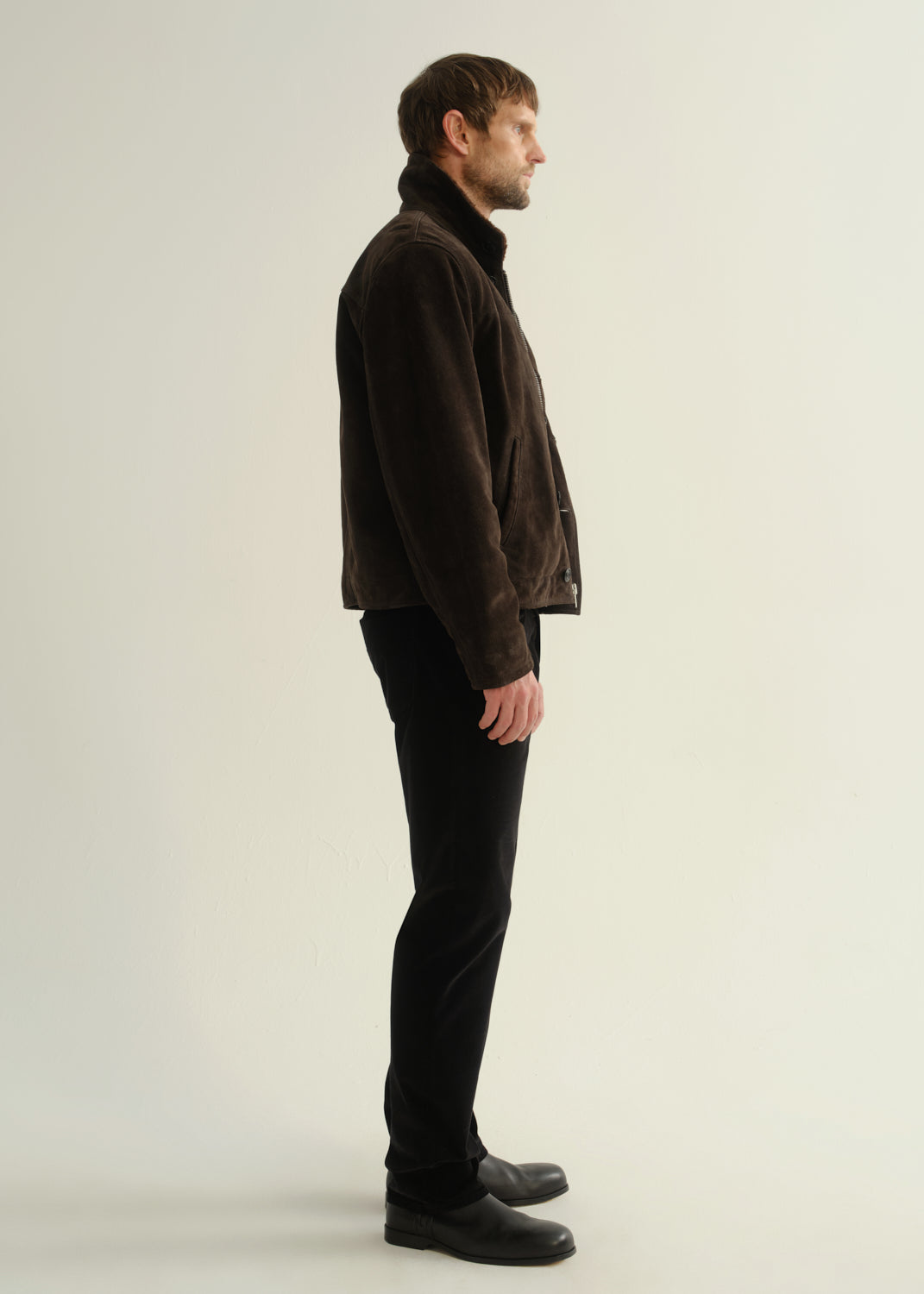Person wearing black straight leg jeans with a dark brown jacket and black boots, side full body view on a light grey background.