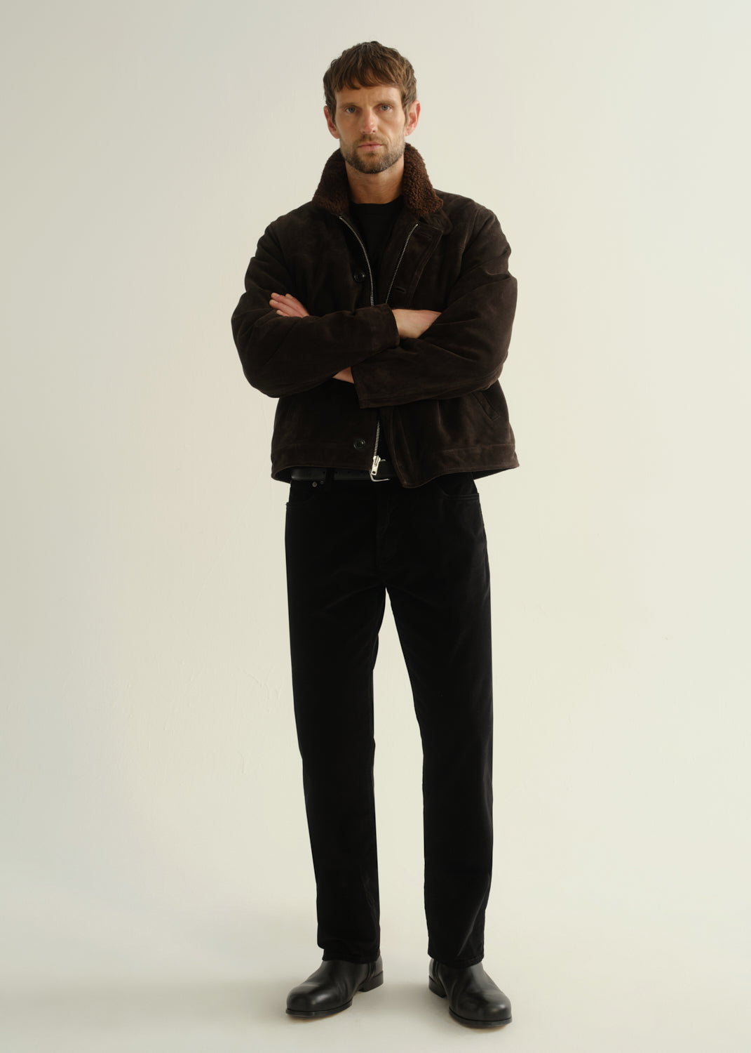 Person wearing black straight leg jeans with a dark brown jacket and black boots, front full body view on a light grey background.