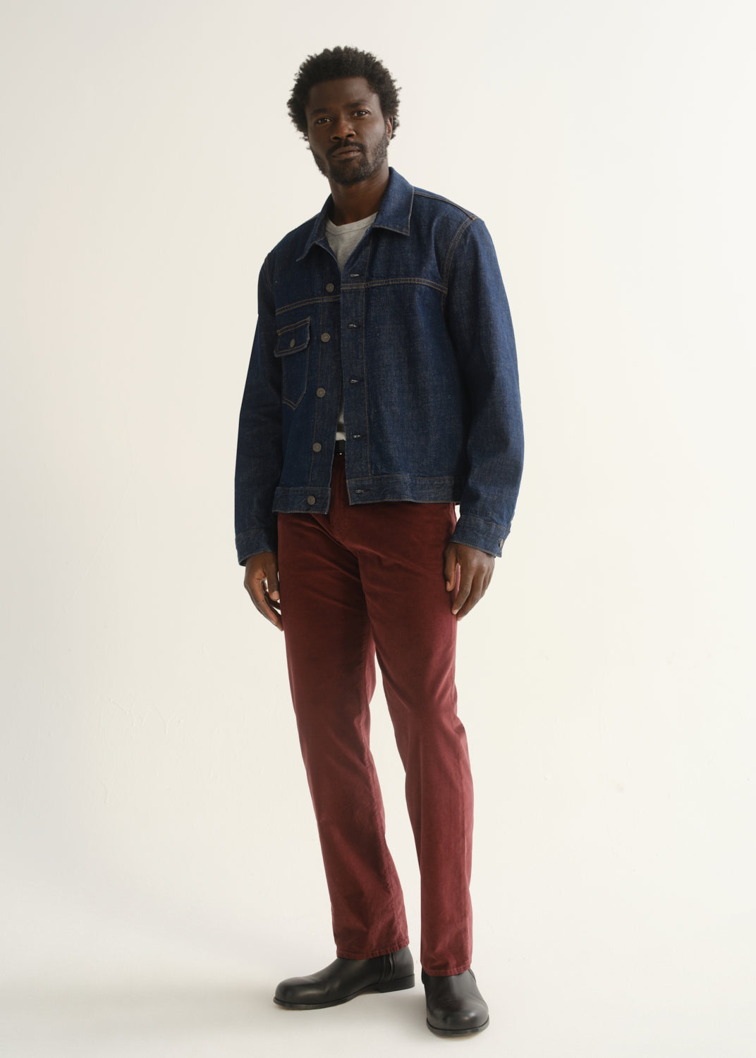 Person wearing burgundy straight leg pants with a medium blue denim jacket, dark shirt, and black shoes, angled front full body view on a light grey background.