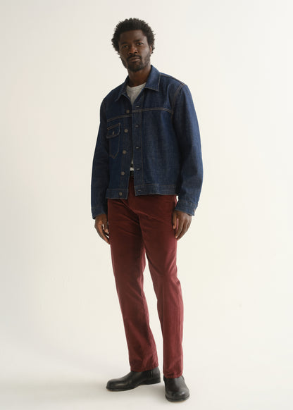 Person wearing burgundy straight leg pants with a medium blue denim jacket, dark shirt, and black shoes, angled front full body view on a light grey background.