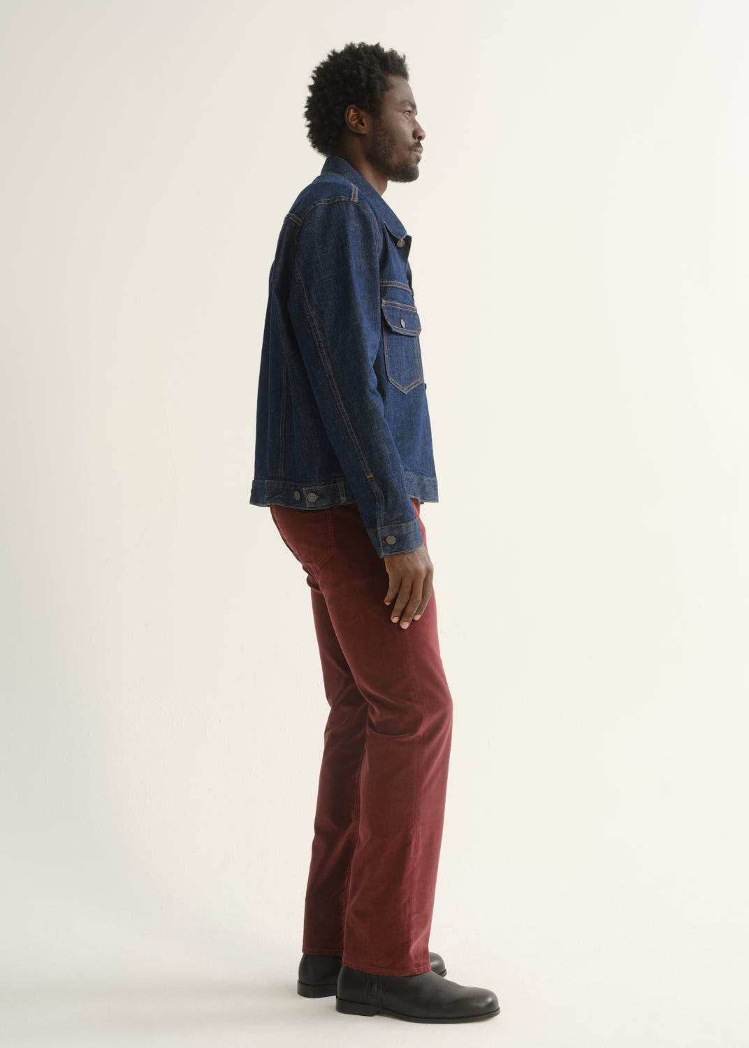 Person wearing burgundy straight leg pants with a medium blue denim jacket and black shoes, side full body view on a light grey background.