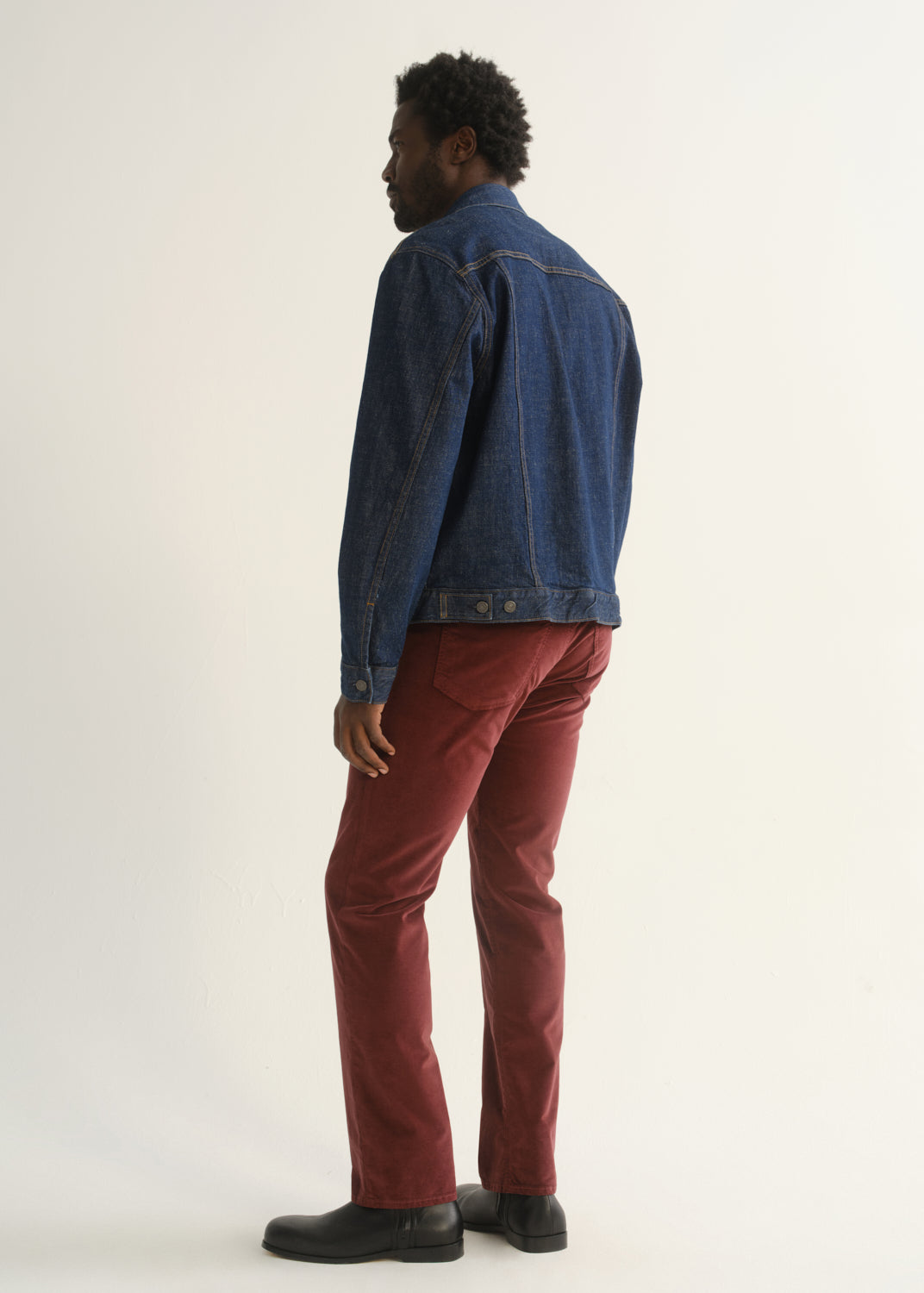 Person wearing burgundy straight leg pants with a medium blue denim jacket and black shoes, back full body view on a light grey background.