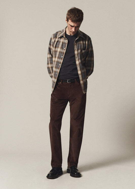 A man wearing a brown and beige plaid flannel shirt over a black t-shirt, paired with dark brown trousers and black leather shoes, standing against a neutral light gray studio background.