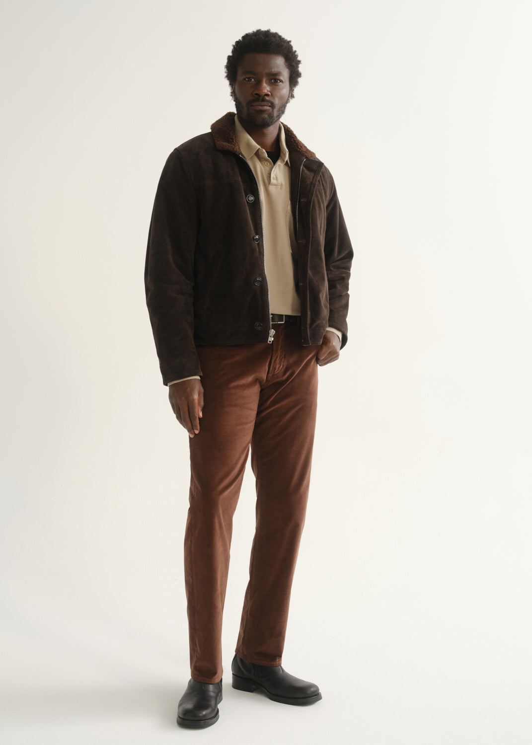 Person wearing rust brown slim straight leg pants with a dark brown jacket, cream shirt, and black shoes, front full body view on a light grey background.
