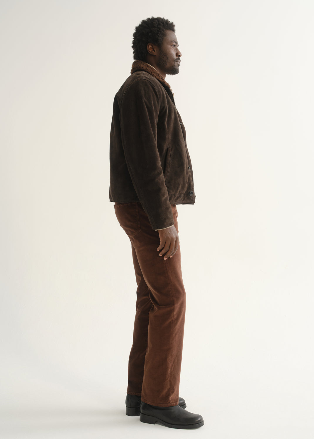 Person wearing rust brown slim straight leg pants with a dark brown jacket and black shoes, side full body view on a light grey background.
