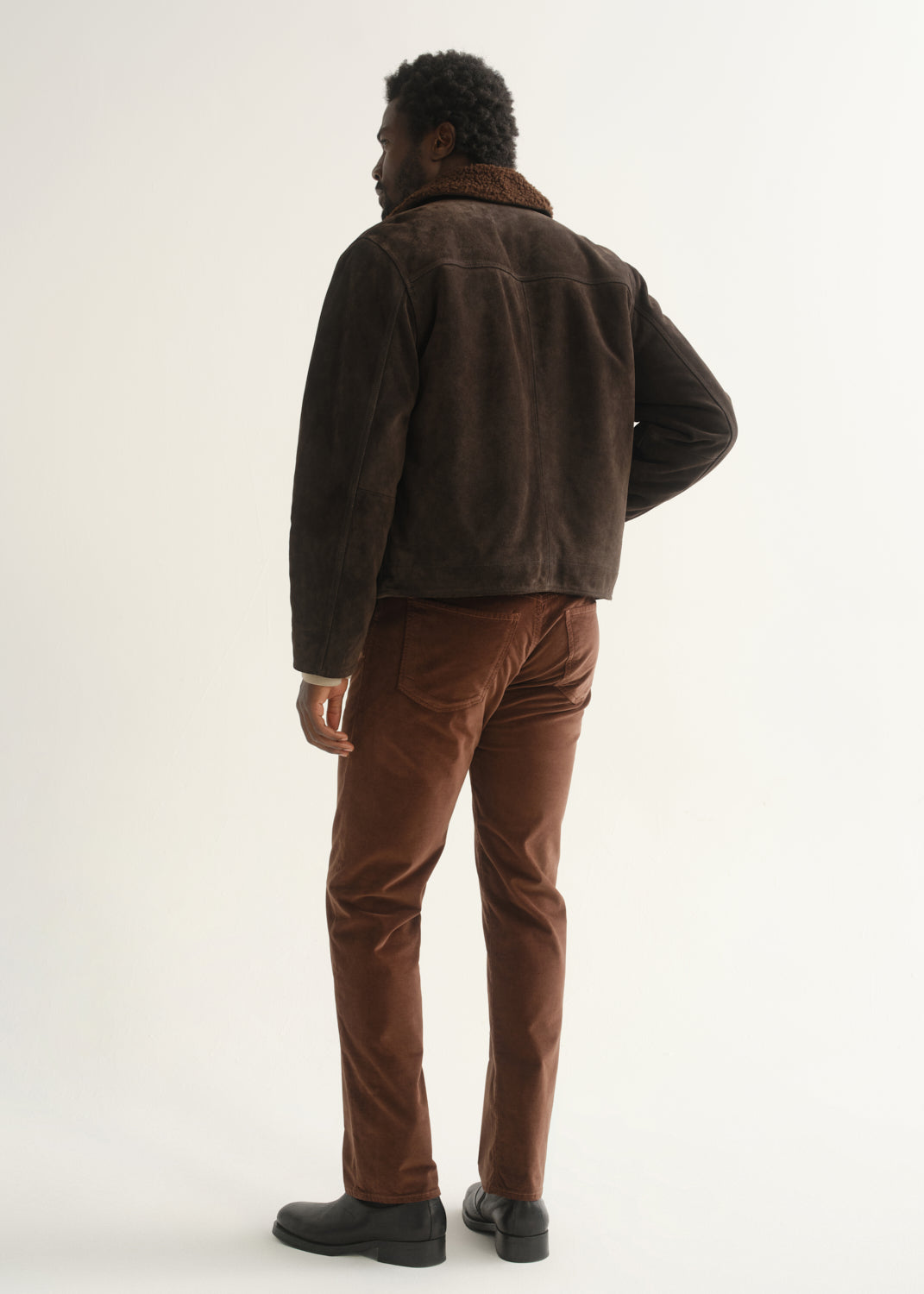 Person wearing rust brown slim straight leg pants with a dark brown jacket and black shoes, back full body view on a light grey background.