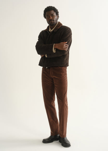 Person wearing rust brown slim straight leg pants with a dark brown jacket and black shoes, angled front full body view with arms crossed on a light grey background.