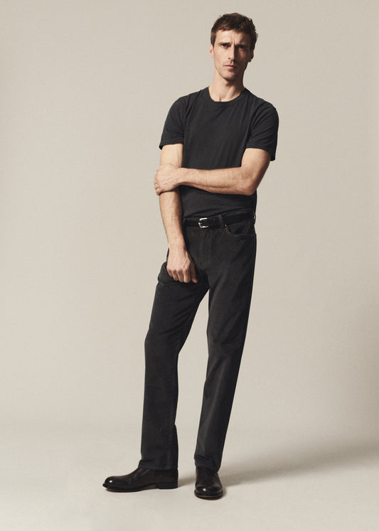 A man modeling a black short-sleeve crewneck t-shirt and black trousers with a black belt, standing against a neutral light beige studio background.