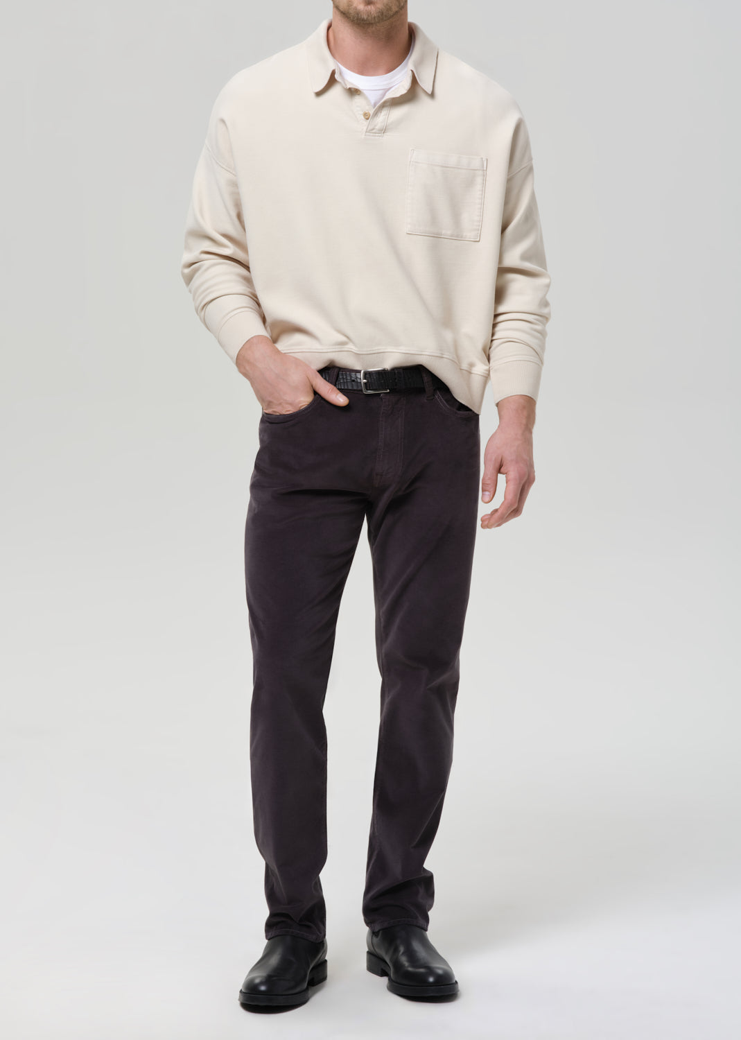 Man wearing a beige sweater and dark pants on a grey background