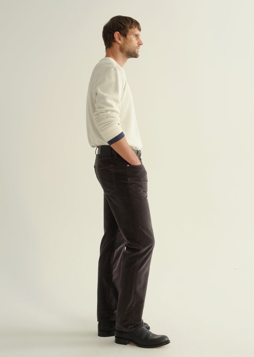 Person wearing dark charcoal straight leg jeans with a cream long sleeve shirt and black boots, side full body view on a light grey background.