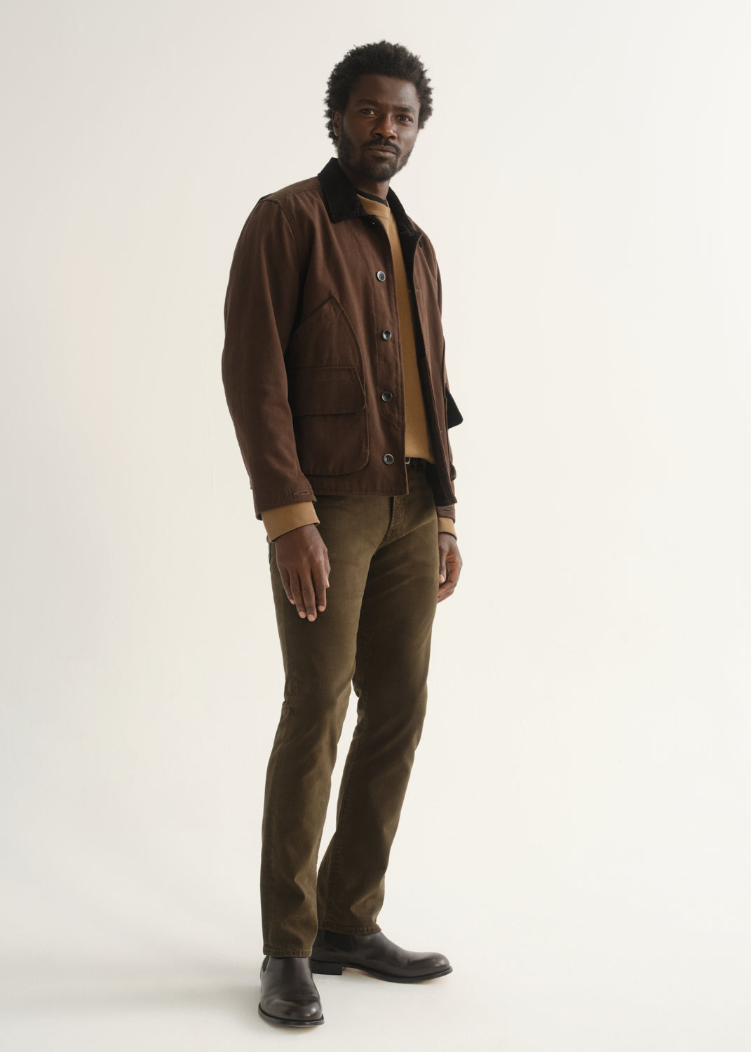 Person wearing dark brown straight leg pants with a brown jacket, dark shirt, and black shoes, angled front full body view on a light grey background.