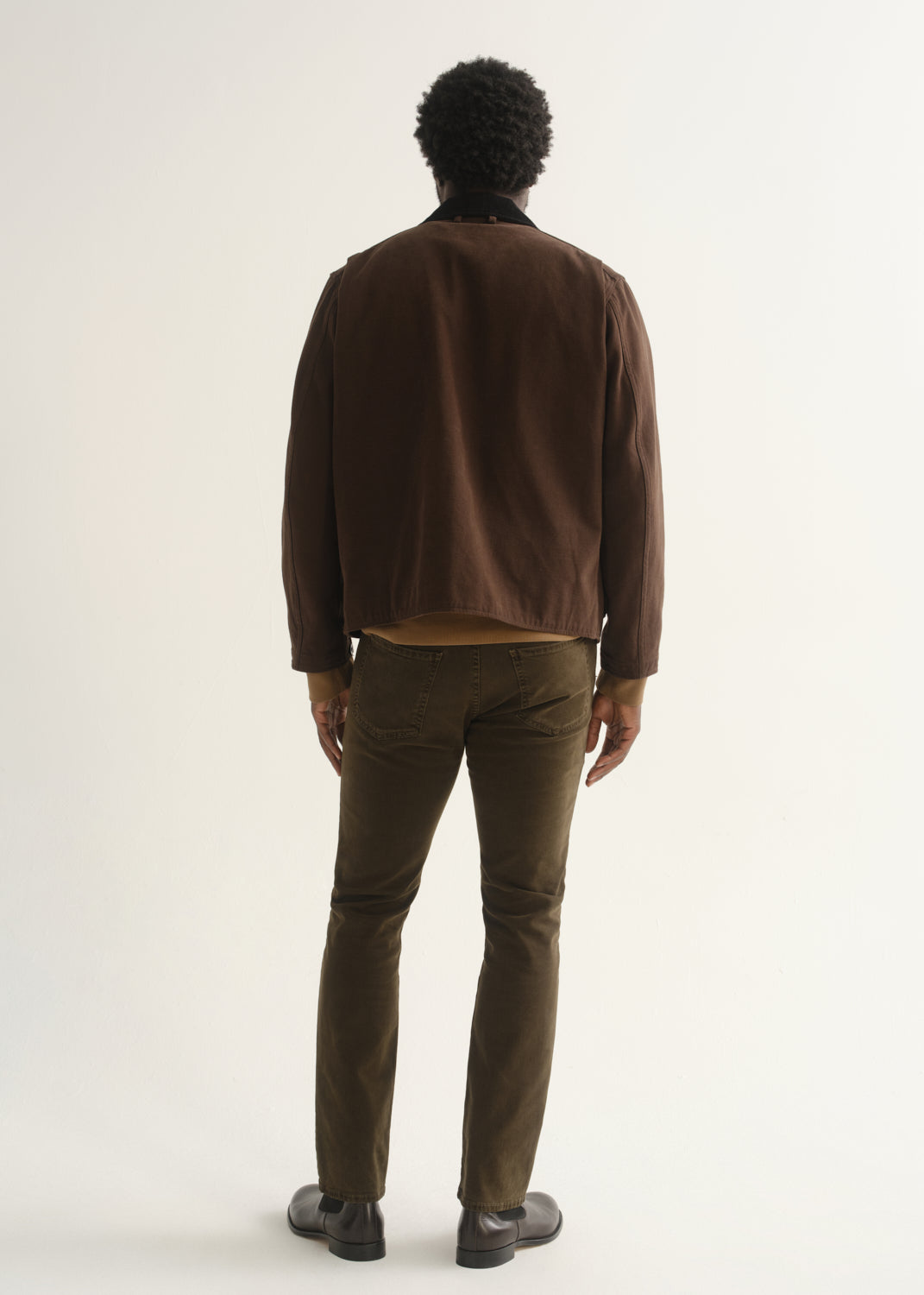 Person wearing dark brown straight leg pants with a brown jacket and black shoes, back full body view on a light grey background.