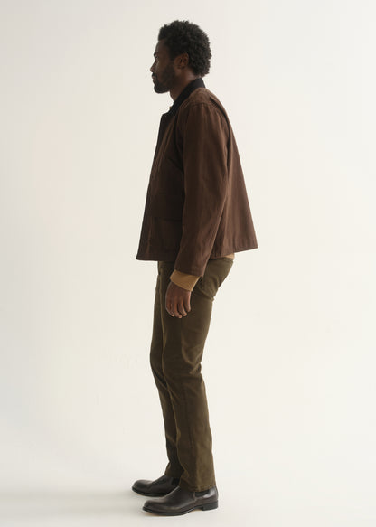 Person wearing dark brown straight leg pants with a brown jacket and black shoes, side full body view on a light grey background.