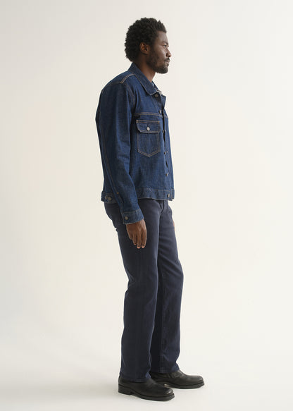 Person wearing dark indigo straight leg jeans with a medium blue denim jacket and black shoes, side full body view on a light grey background.
