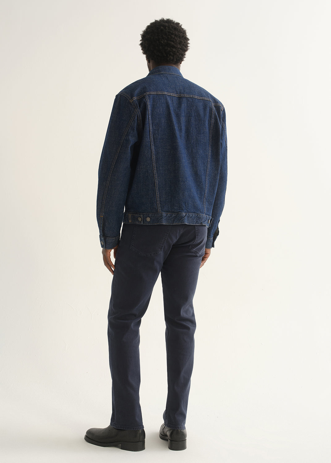 Person wearing dark indigo straight leg jeans with a medium blue denim jacket and black shoes, back full body view on a light grey background.
