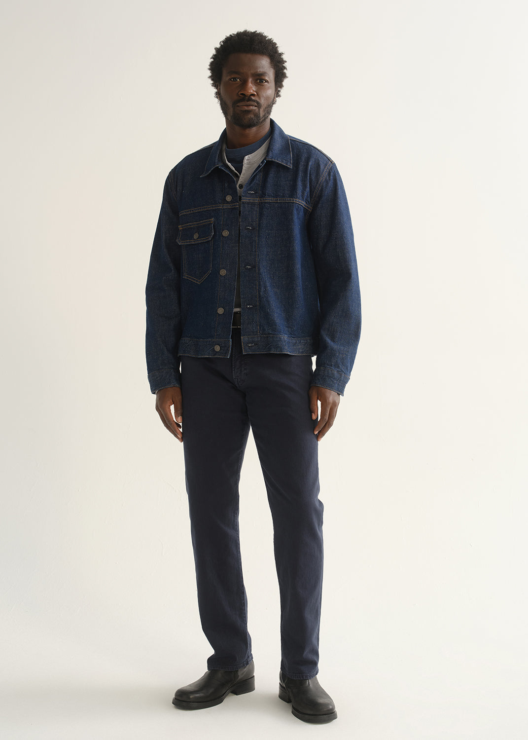 Person wearing dark indigo straight leg jeans with a medium blue denim jacket, dark shirt, and black shoes, front full body view on a light grey background.