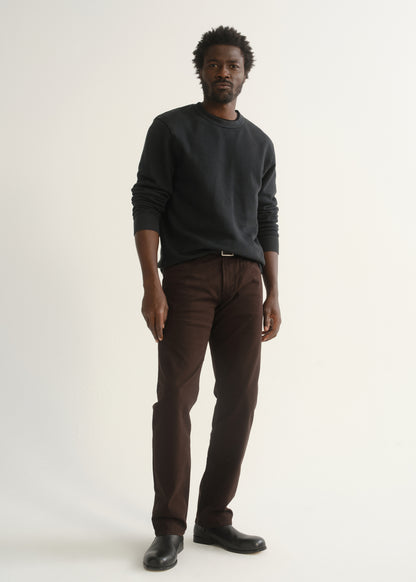 Person wearing dark brown straight leg pants with a dark charcoal long sleeve crewneck sweater and black shoes, angled front full body view on a light grey background.