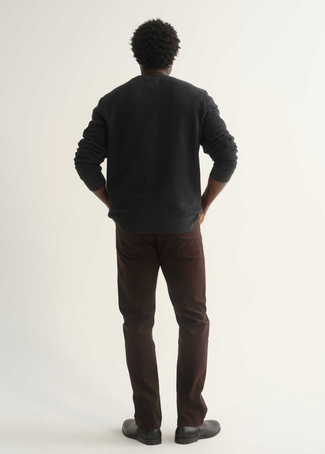 Person wearing dark brown straight leg pants with a dark charcoal long sleeve crewneck sweater and black shoes, back full body view on a light grey background.