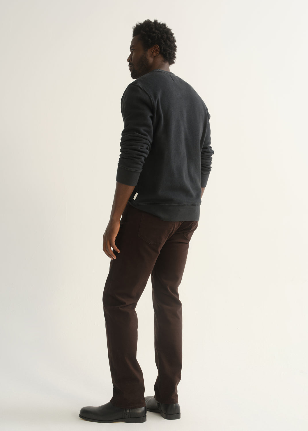 Person wearing dark brown straight leg pants with a dark charcoal long sleeve crewneck sweater and black shoes, side full body view on a light grey background.