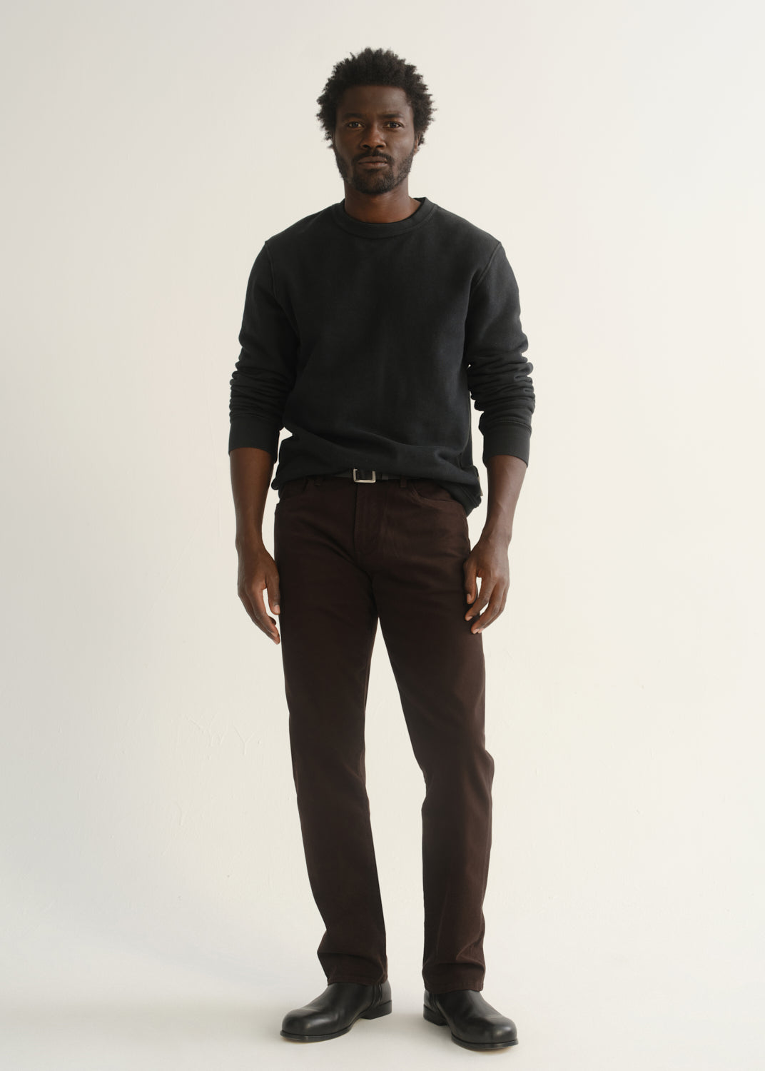 Person wearing dark brown straight leg pants with a dark charcoal long sleeve crewneck sweater and black shoes, front full body view on a light grey background.