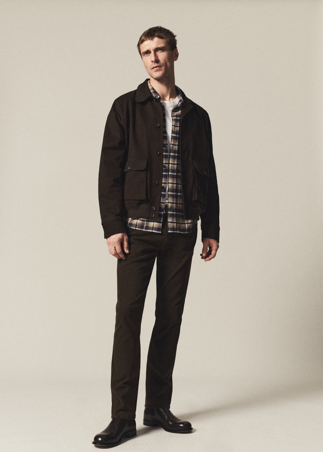 A man modeling a dark brown utility jacket over a plaid button-down shirt and dark brown trousers, standing against a plain light beige studio background.