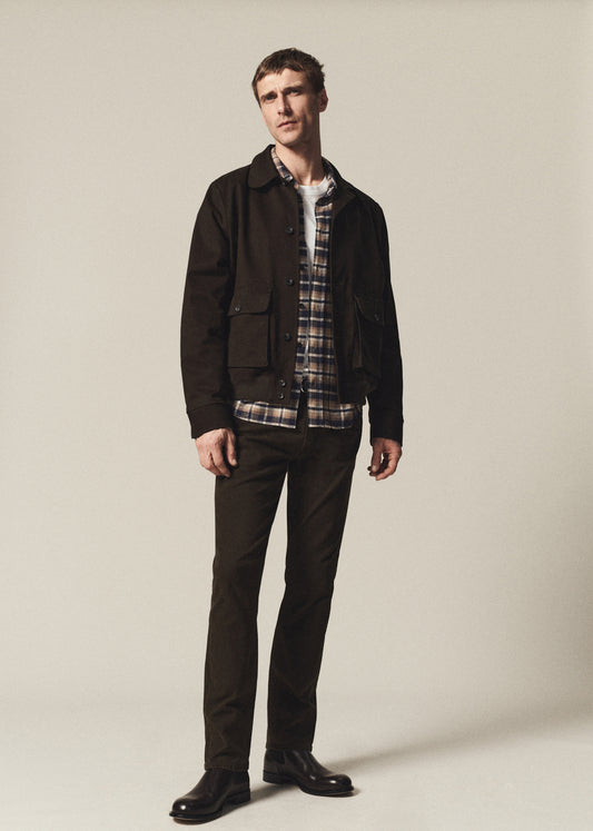 A man modeling a dark brown utility jacket over a plaid button-down shirt and dark brown trousers, standing against a plain light beige studio background.