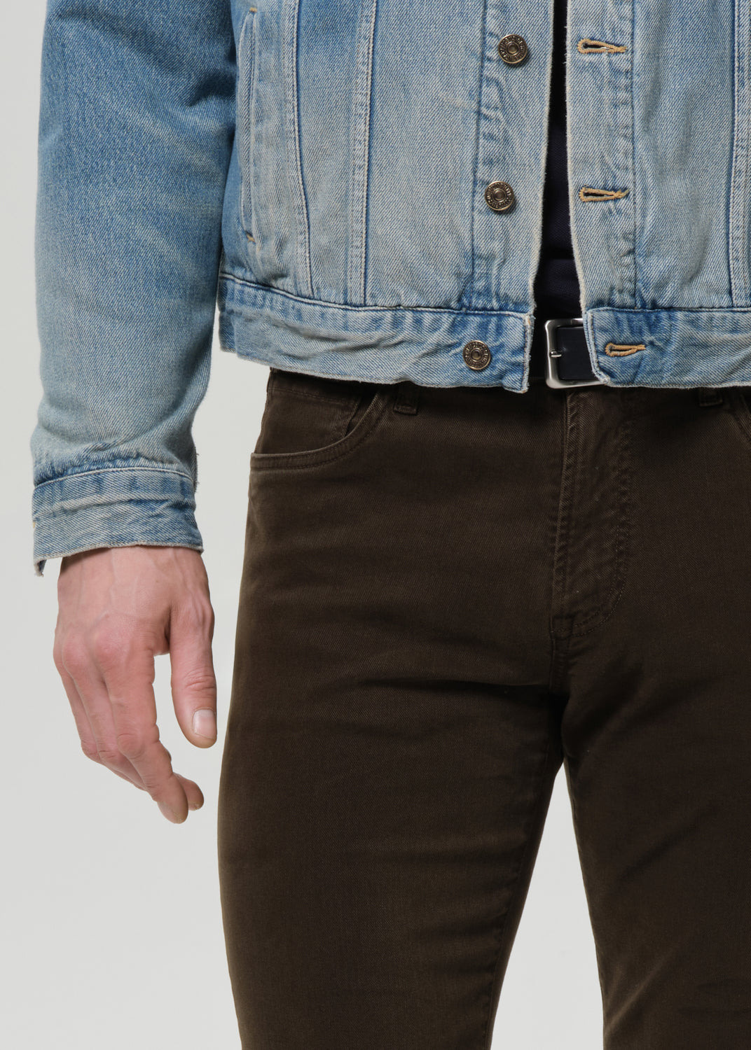 Close-up of a person wearing a light blue denim jacket and dark brown pants against a grey background.