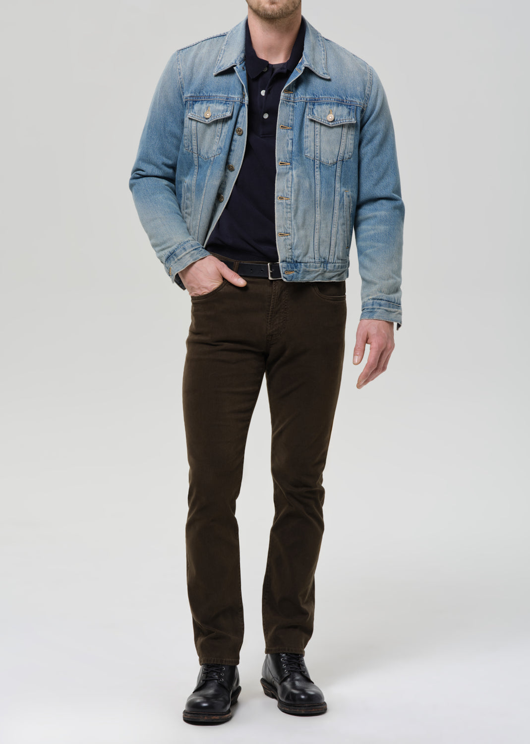 Person wearing a denim jacket, black shirt, and brown pants on a grey background.