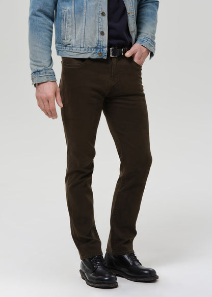 A person wearing slim-fit dark brown trousers, styled with a light blue denim jacket, black shirt, and black leather shoes, standing against a grey background.