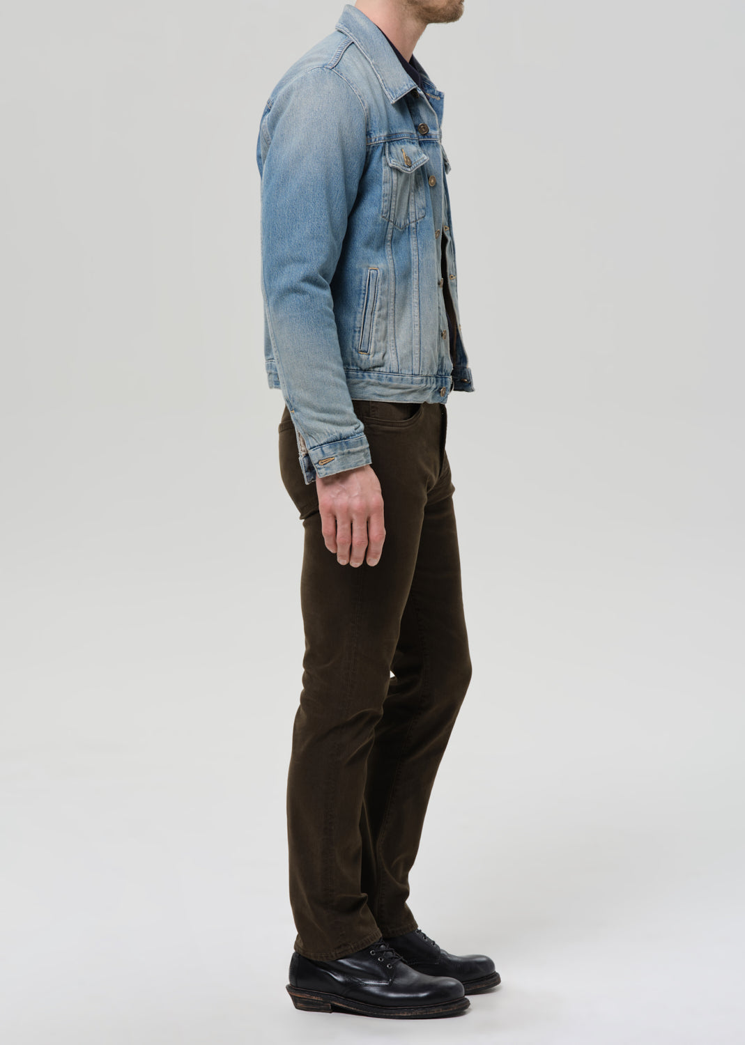 A man shown in profile wearing a light blue denim jacket, dark brown trousers, and black leather shoes against a grey background.