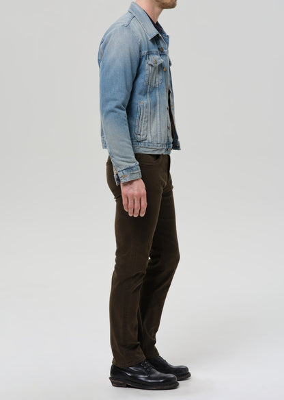 A man shown in profile wearing a light blue denim jacket, dark brown trousers, and black leather shoes against a grey background.