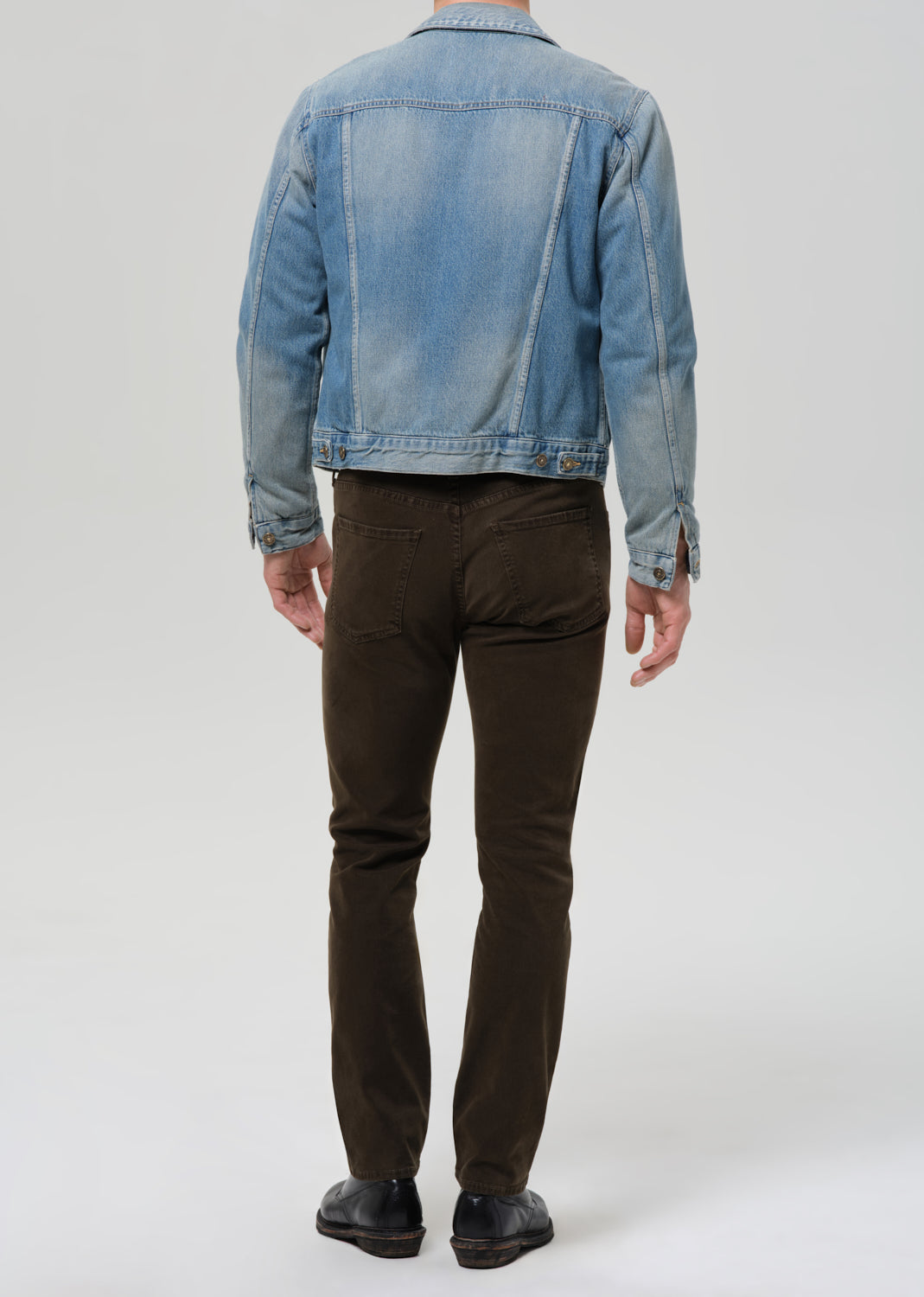 A man viewed from the back wearing a light blue denim jacket, dark brown slim-fit pants, and black leather shoes, standing against a grey background.